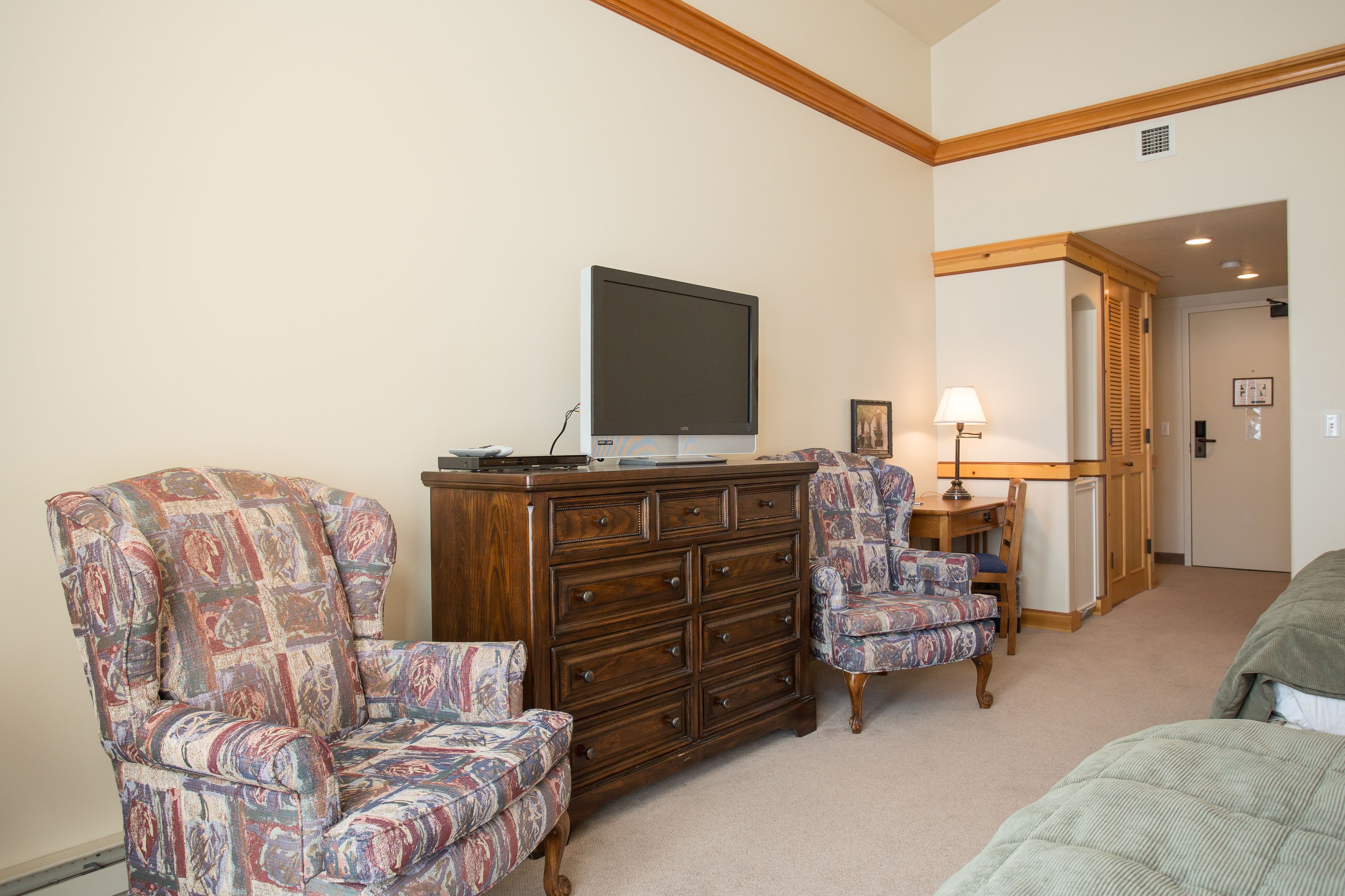 A room with a large wooden dresser, two patterned armchairs, a television on top, a lamp, and a bed.