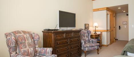 A room with a large wooden dresser, two patterned armchairs, a television on top, a lamp, and a bed.