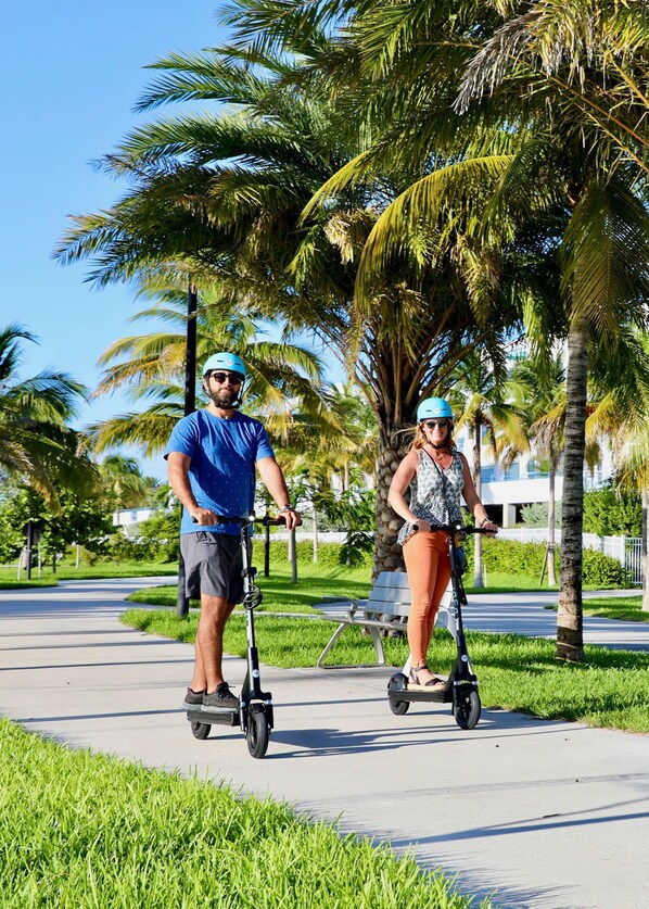 Two people riding electric scooters on a paved path with palm trees and a bench in the background.