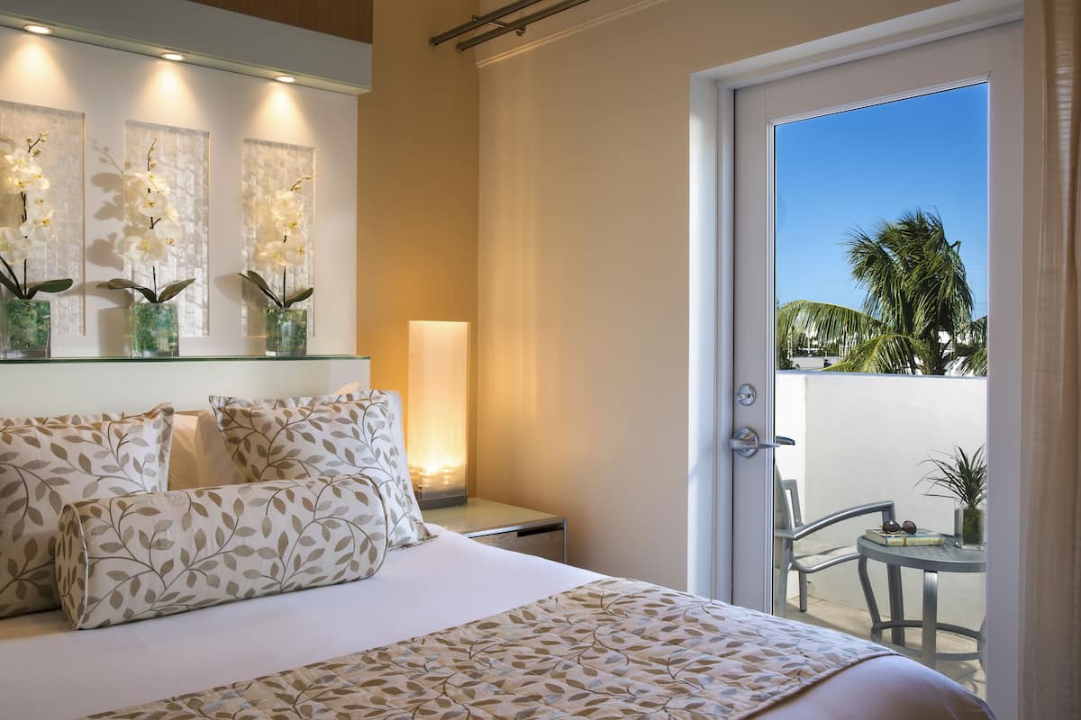Quarto de hotel com cama, mesa de cabeceira, abajur, espelho e vista para a varanda.