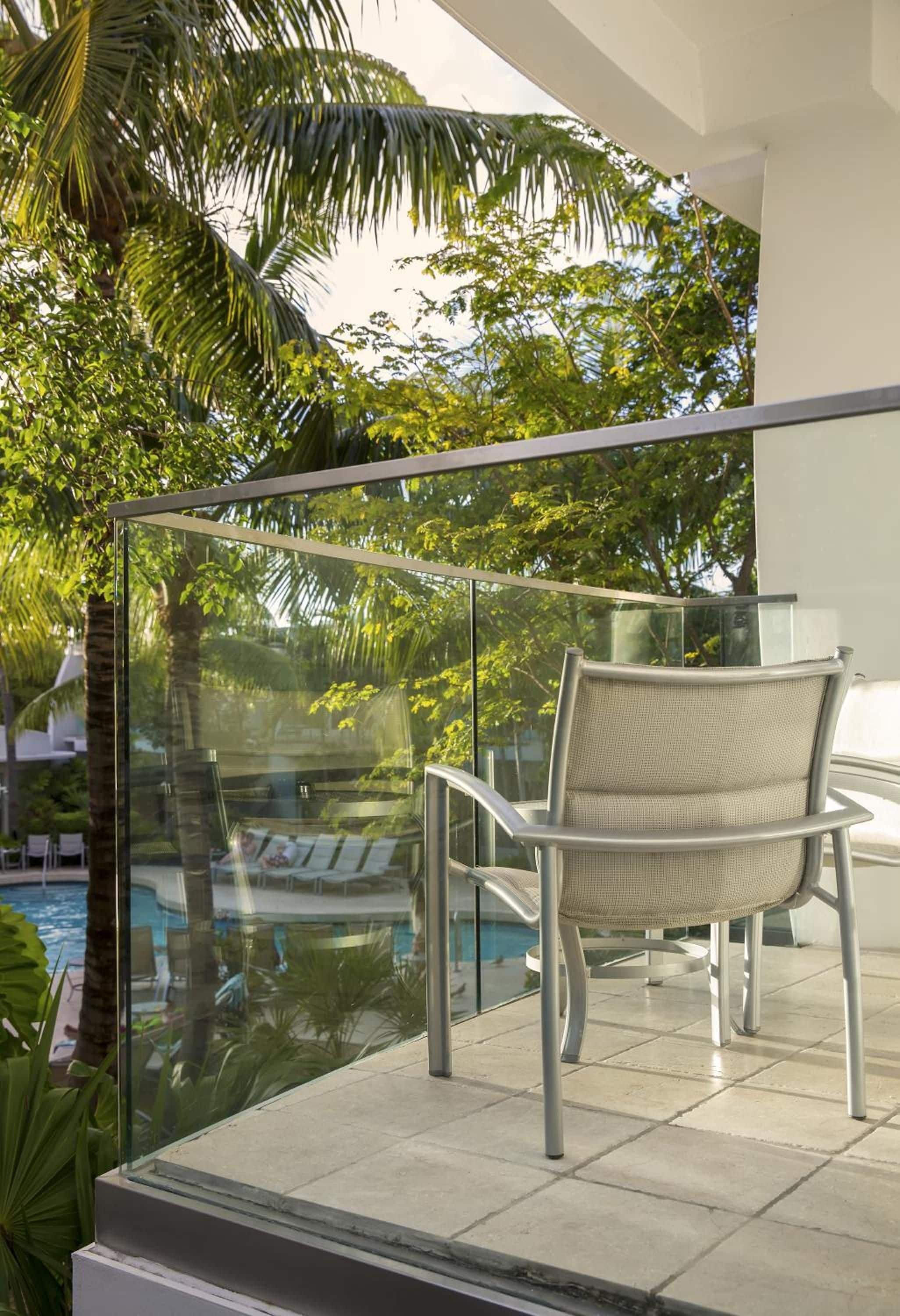 Un balcón con barandilla de vidrio, una silla de mimbre y vista a una piscina y palmeras.