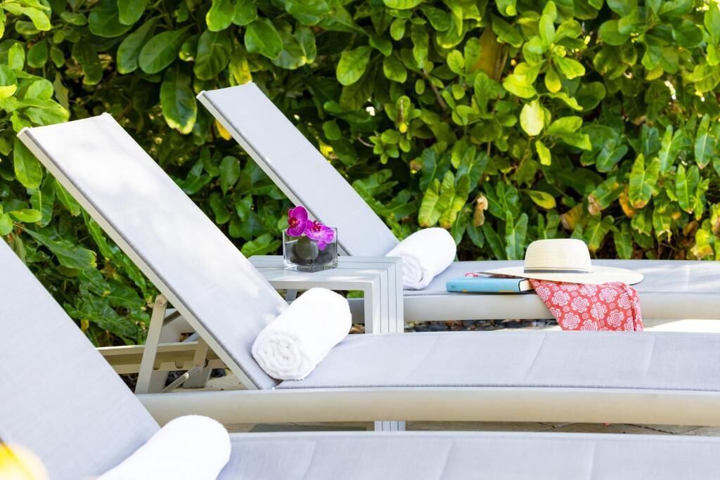 Un espace aménagé au bord de la piscine, avec des chaises longues, une petite table ornée d’un vase et d’un chapeau, et une haie verdoyante en arrière-plan.