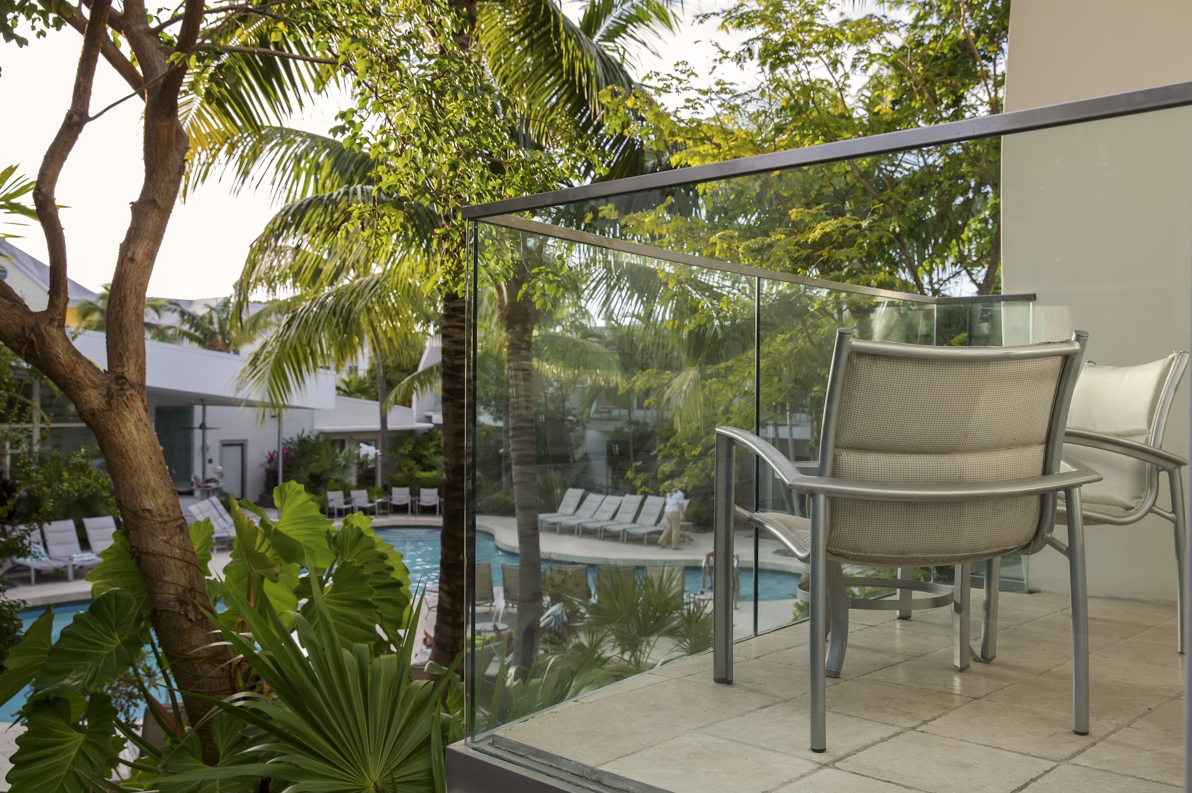 Ein Balkon mit Glasgeländer, einem Stuhl und Blick auf einen Poolbereich mit Liegestühlen und Palmen.