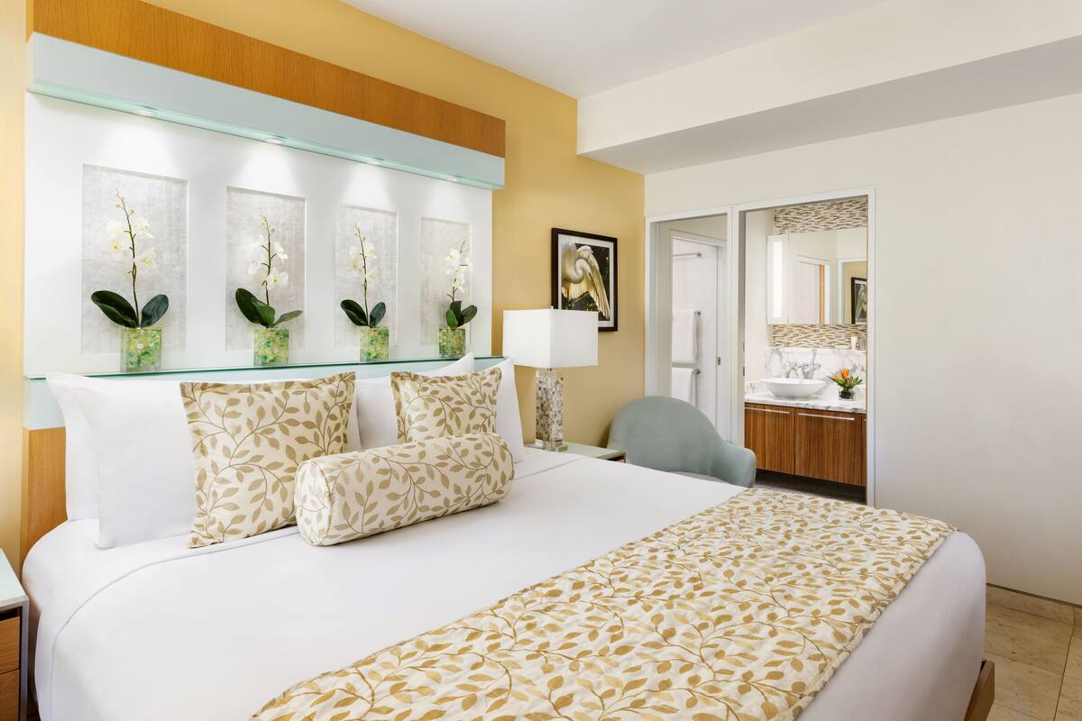 Quarto de hotel com uma cama grande, um painel de vidro com vasos de orquídeas, uma escrivaninha com abajur, uma cadeira e um banheiro visível por uma porta aberta.