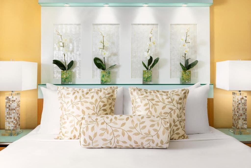 Habitación de hotel con cama, dos mesitas de noche y un separador de vidrio con tres jarrones de orquídeas.