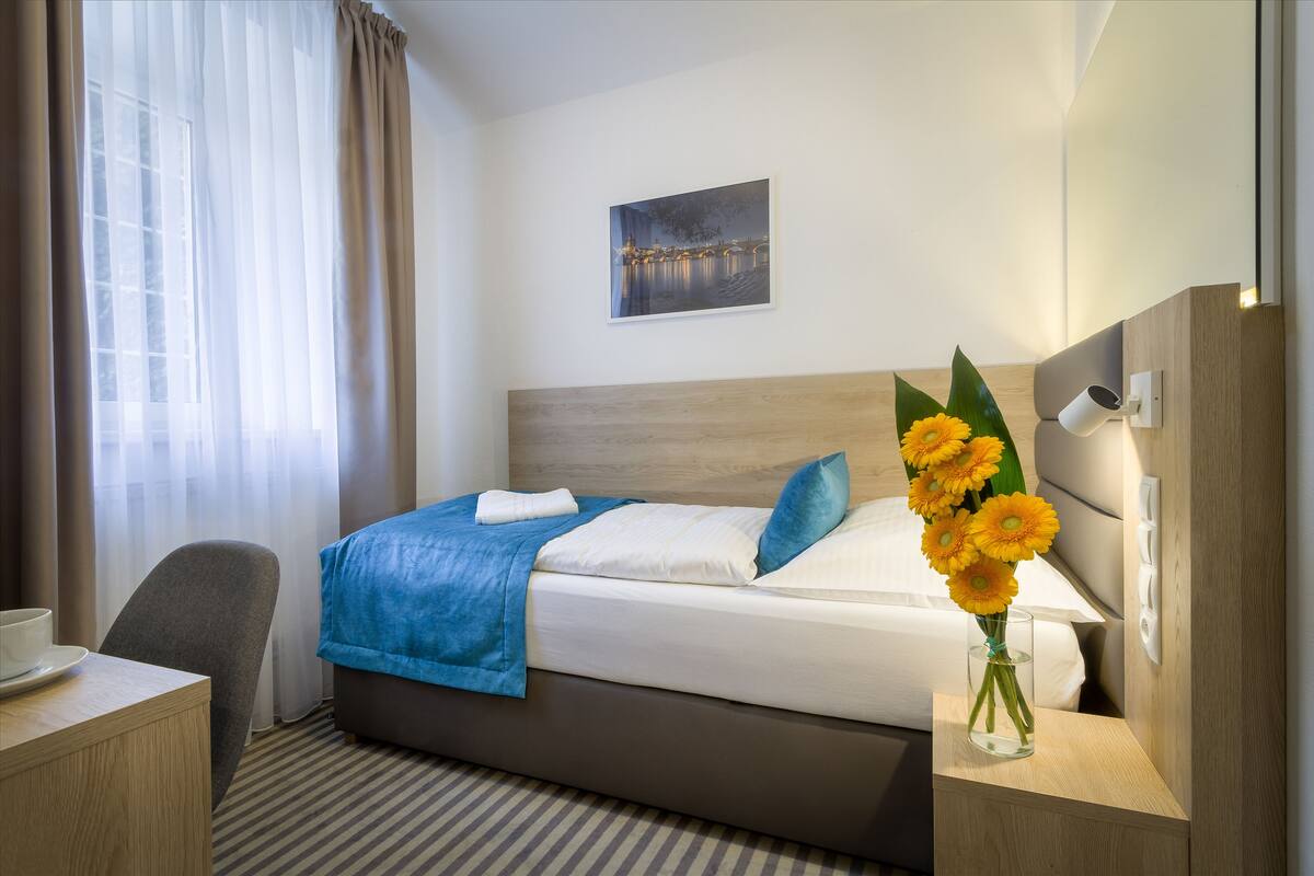 A hotel room with a bed, a desk, a chair, a vase with flowers, and a painting on the wall.