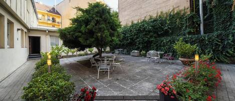 Un patio con un árbol en el centro, mobiliario exterior y plantas en macetas.