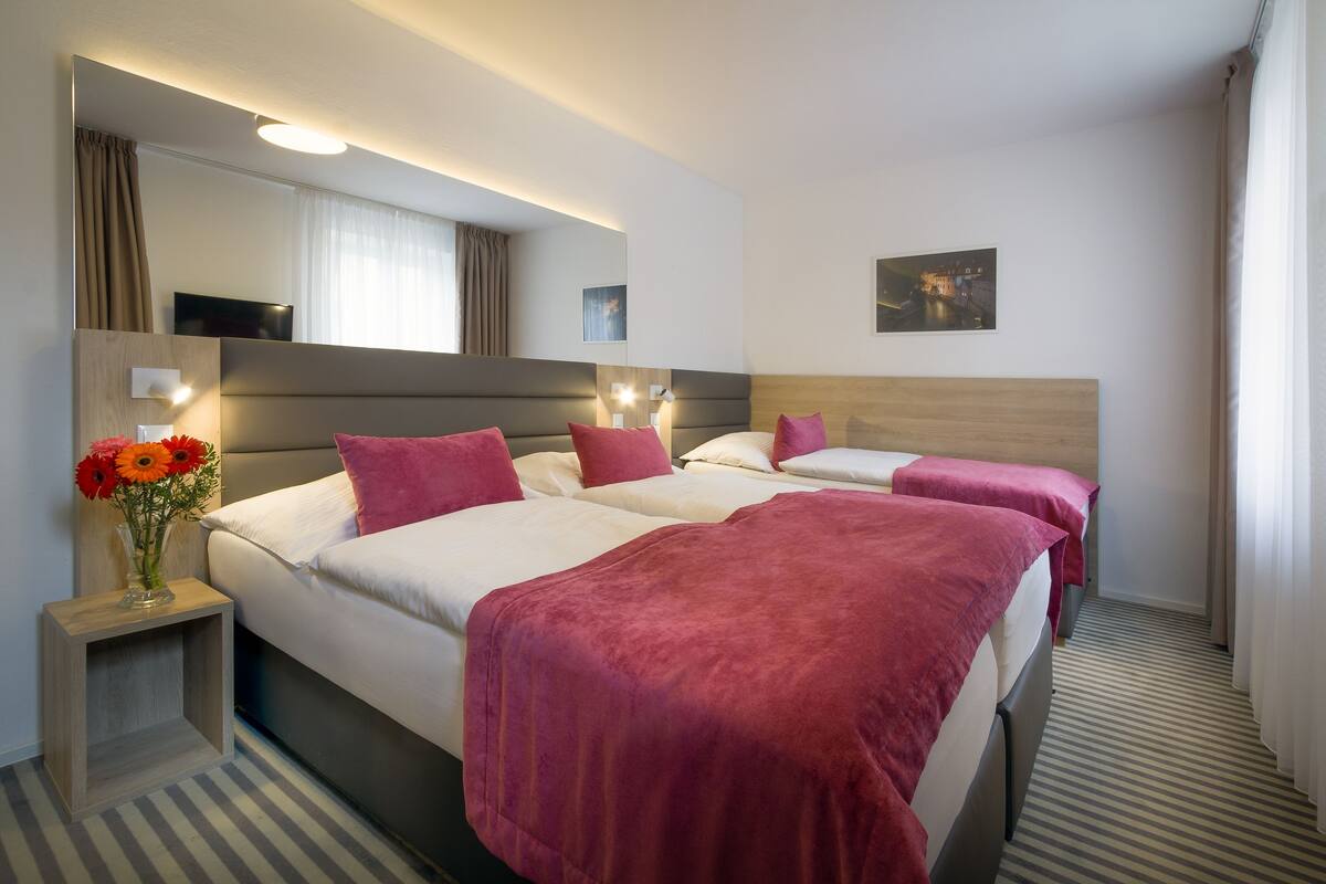 A hotel room with two beds, a bedside table with a vase of flowers, a flat-screen TV, and a large mirror.