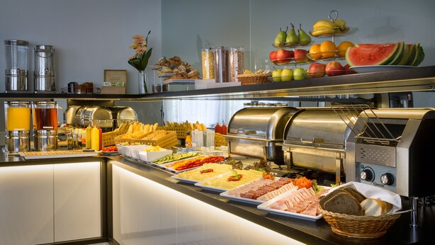 Un bufé con distintos alimentos, como frutas, verduras y carnes, junto con una estación de café y una cesta de pan.