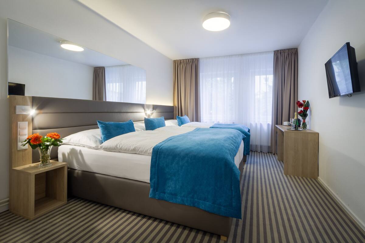 A hotel room with a large bed, a bedside table with a vase of flowers, a flat-screen TV mounted on the wall, and a large mirror above the bed.