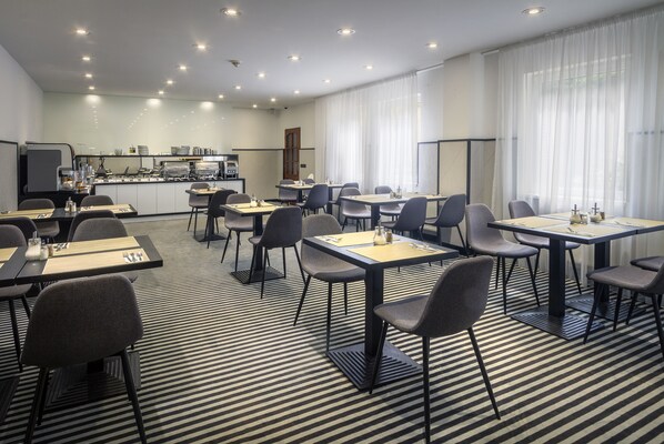 A modern hotel dining area with multiple tables and chairs, a kitchen in the background, and large windows with sheer curtains. - Hotel White Lion (Prague)