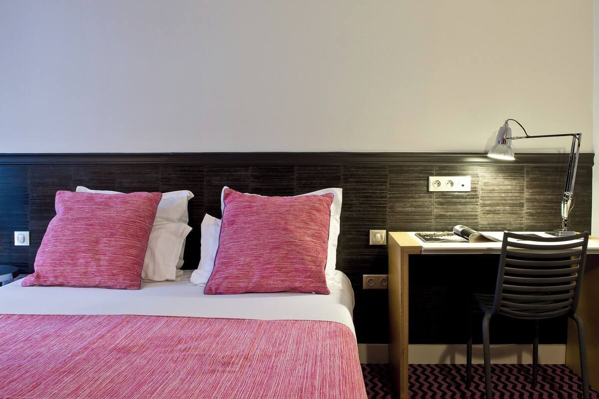 A hotel room with a bed, a desk, a chair, and a lamp.