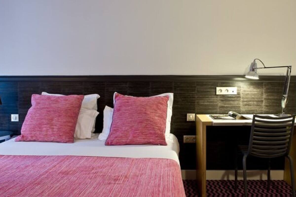 A hotel room with a bed, two pillows, a desk, a chair, a lamp, and a wall-mounted outlet.