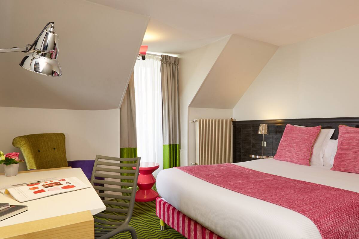 A modern hotel room with a large bed, a desk, a chair, and a lamp.