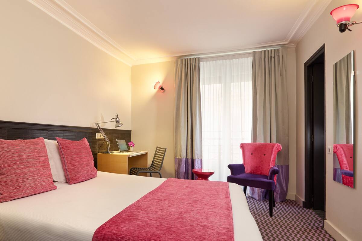 A hotel room with a large bed, a desk with a chair, a small table, and a chair with a red cushion.