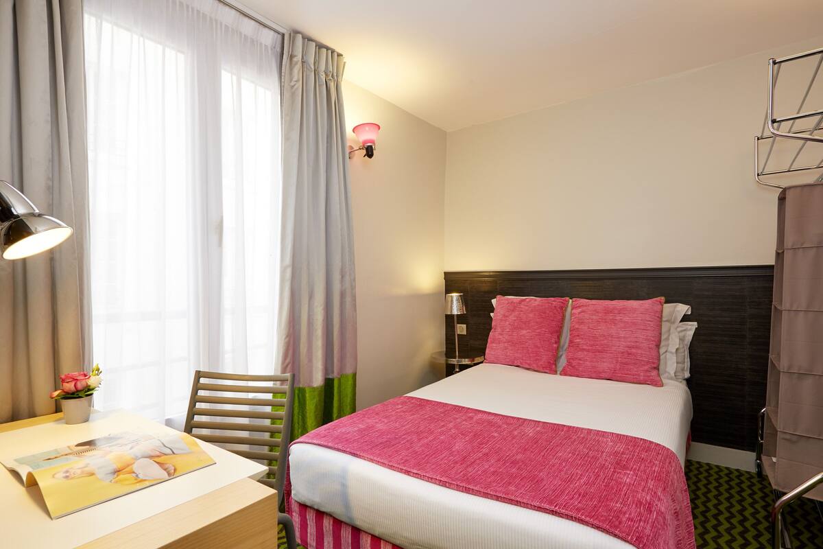 A hotel room with a bed, a desk with a lamp, a chair, a nightstand, and a window with curtains.