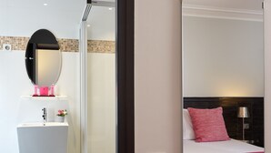 A modern hotel room with a glass shower enclosure, a white sink with a pink soap dispenser, a round mirror, a bed with a black headboard, and a red throw blanket. - Hôtel Antin Trinité (Paris)