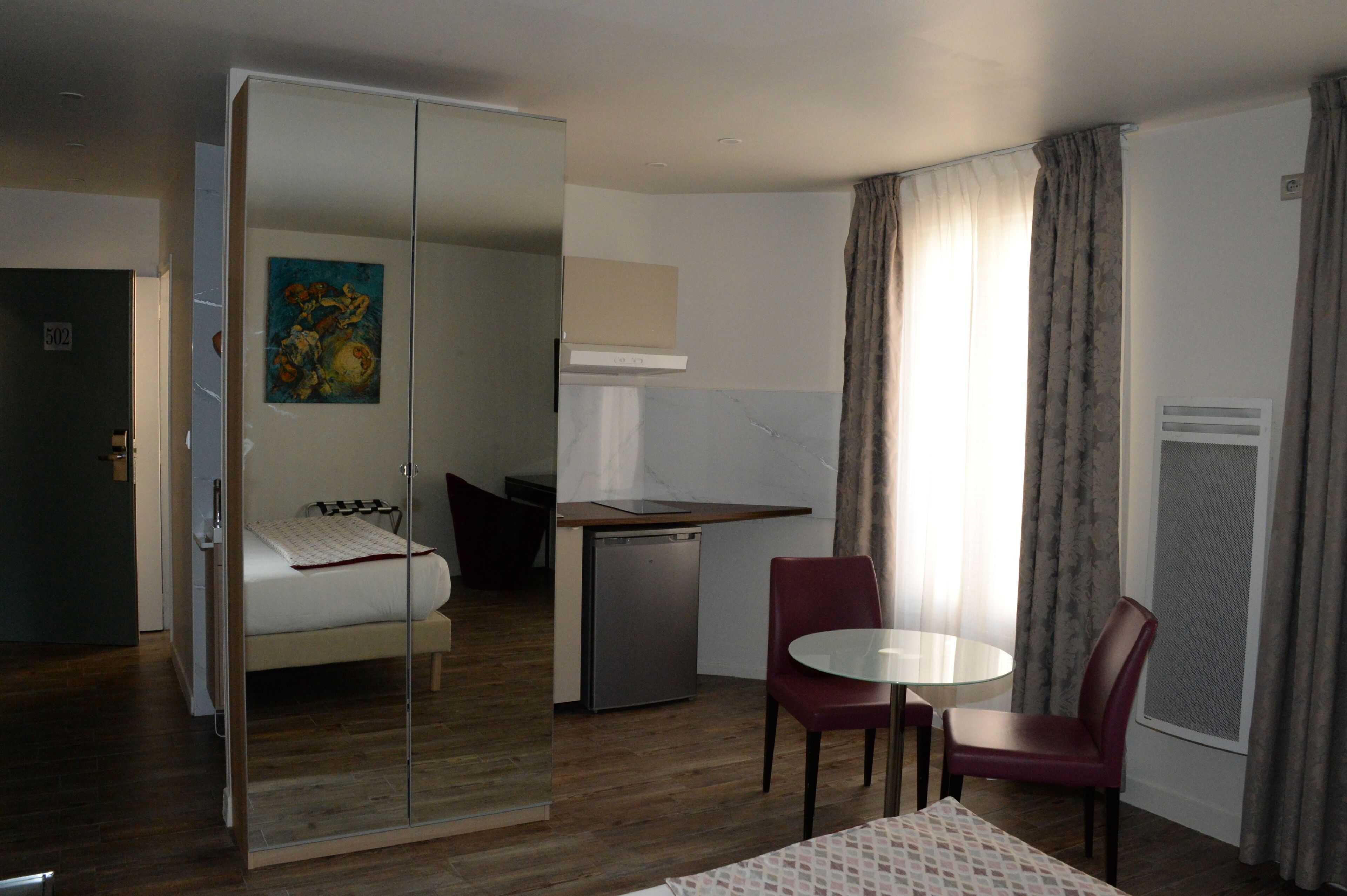 A hotel room with a bed, a small kitchenette, a dining table with two chairs, and a sliding door leading to another room.
