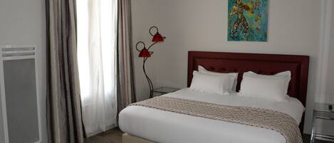 A neatly made bed with a headboard, a window with curtains, a wall-mounted artwork, and a bedside table.