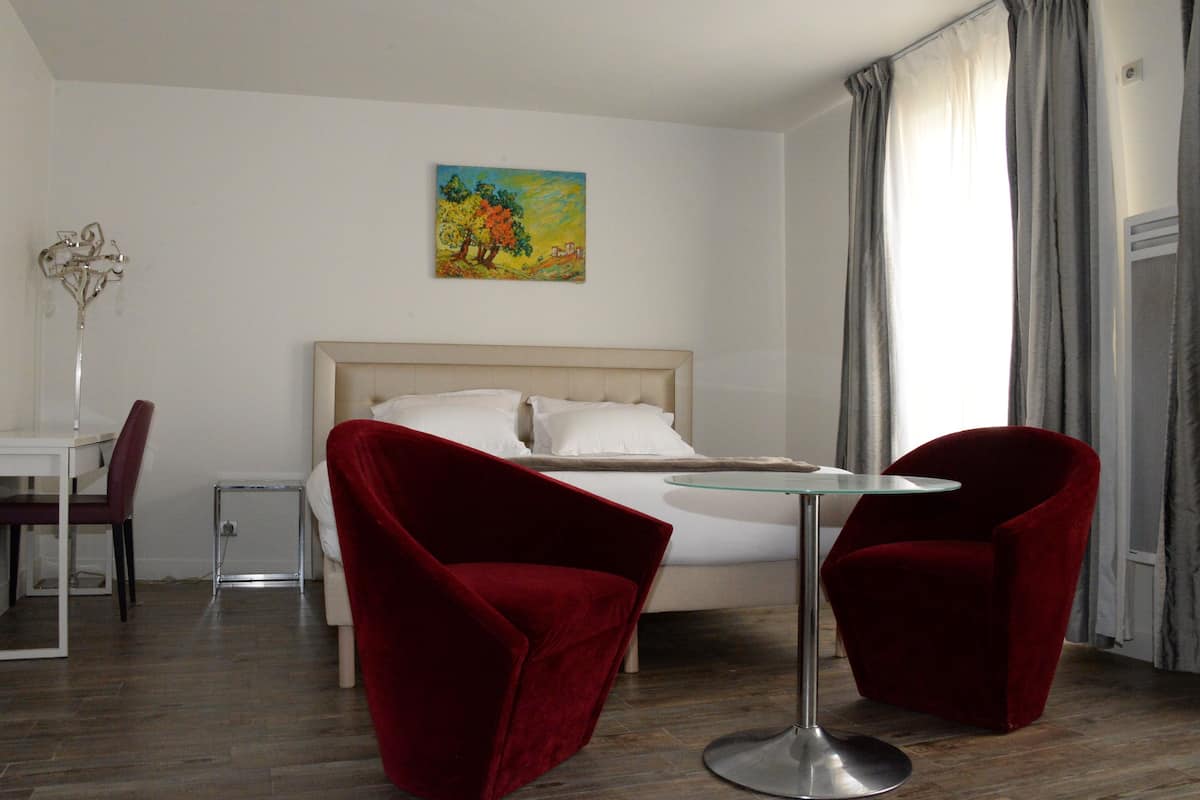 Una camera d'albergo con un letto, due poltrone rosse, un tavolino, un quadro appeso al muro e una finestra con tende.