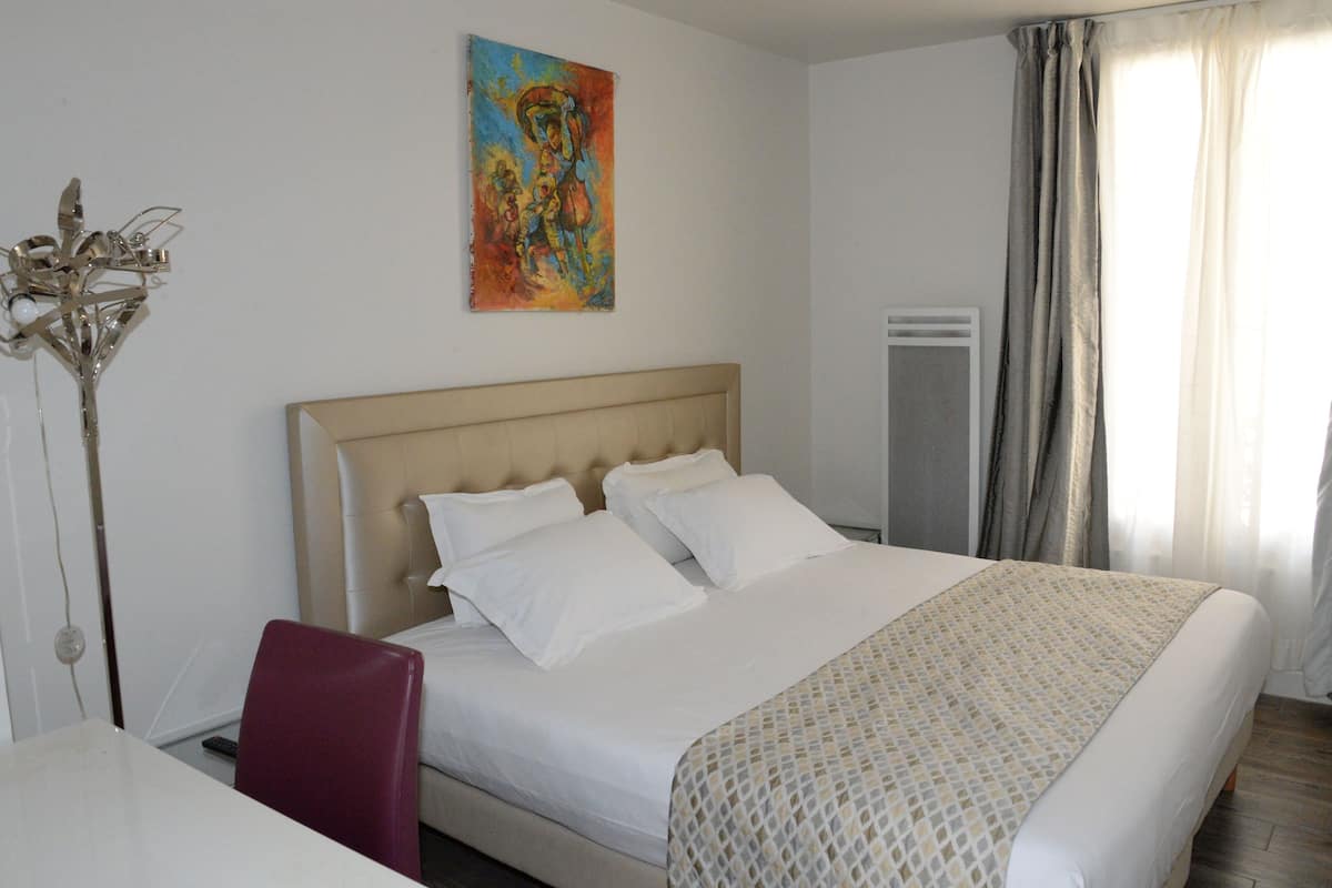 Una camera d'albergo con un letto, una sedia, un quadro appeso e una finestra con tende.