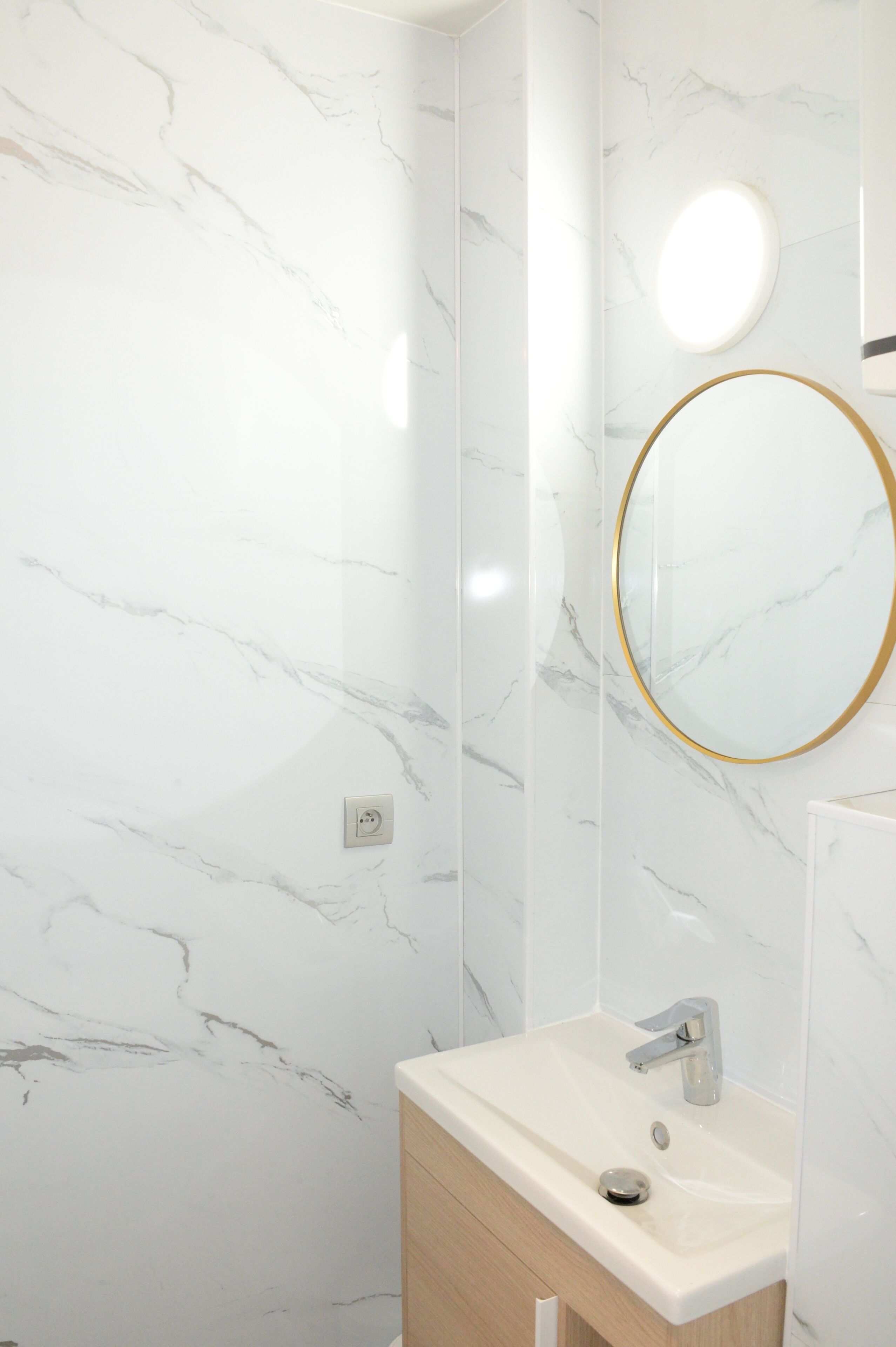 A bathroom with a marble wall, a round mirror with a gold frame, and a modern sink.