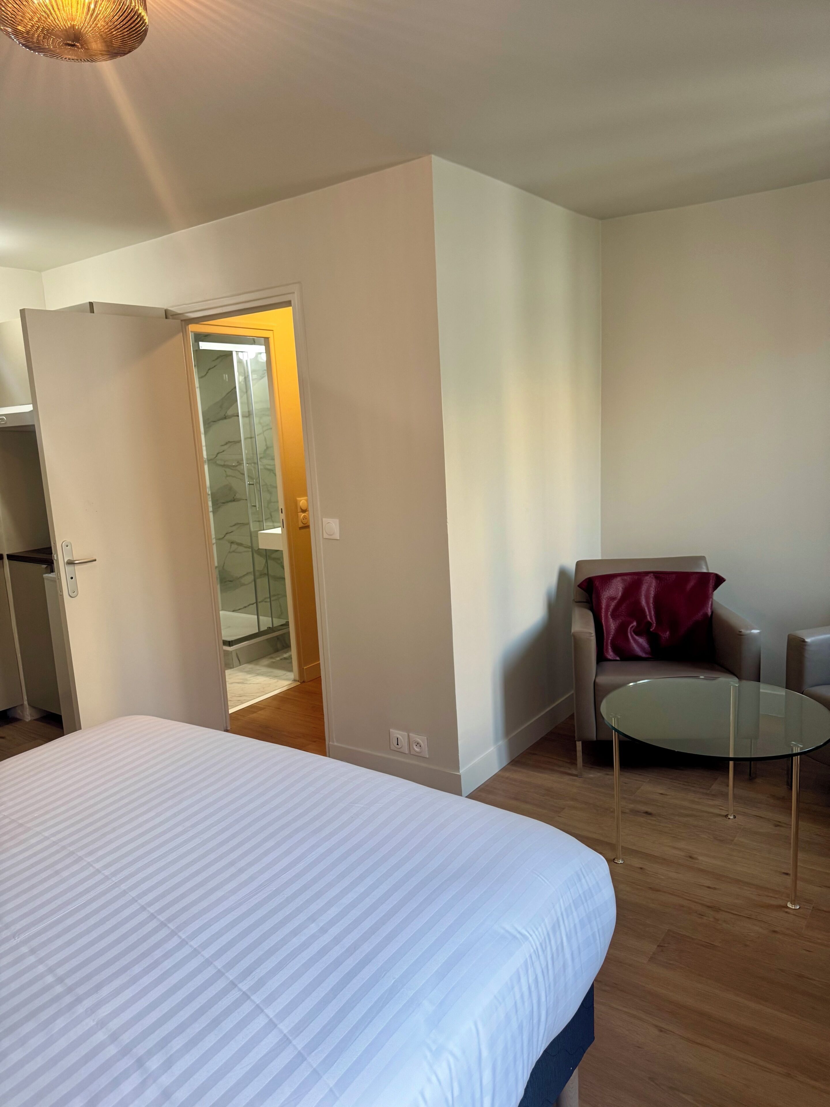 A bedroom with a bed, a glass table, a chair, and a bathroom visible through an open door.