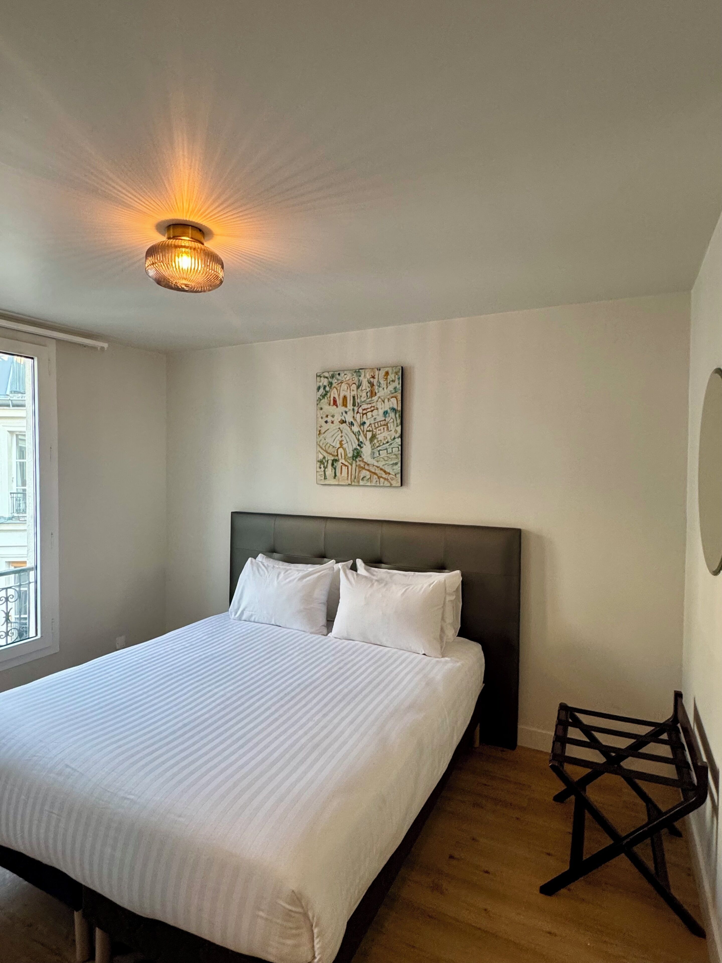 Un dormitorio con cama, cabecera, una obra de arte en la pared, ventana y suelo de madera.