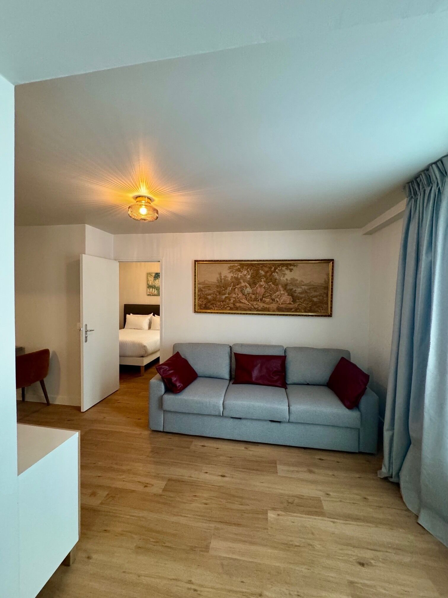 A room with a light blue sofa, a painting on the wall, a bed in the background, and a door leading to another room.