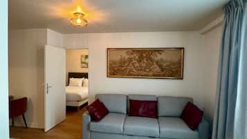 A room with a light blue sofa, a painting on the wall, a bed in the background, and a door leading to another room.