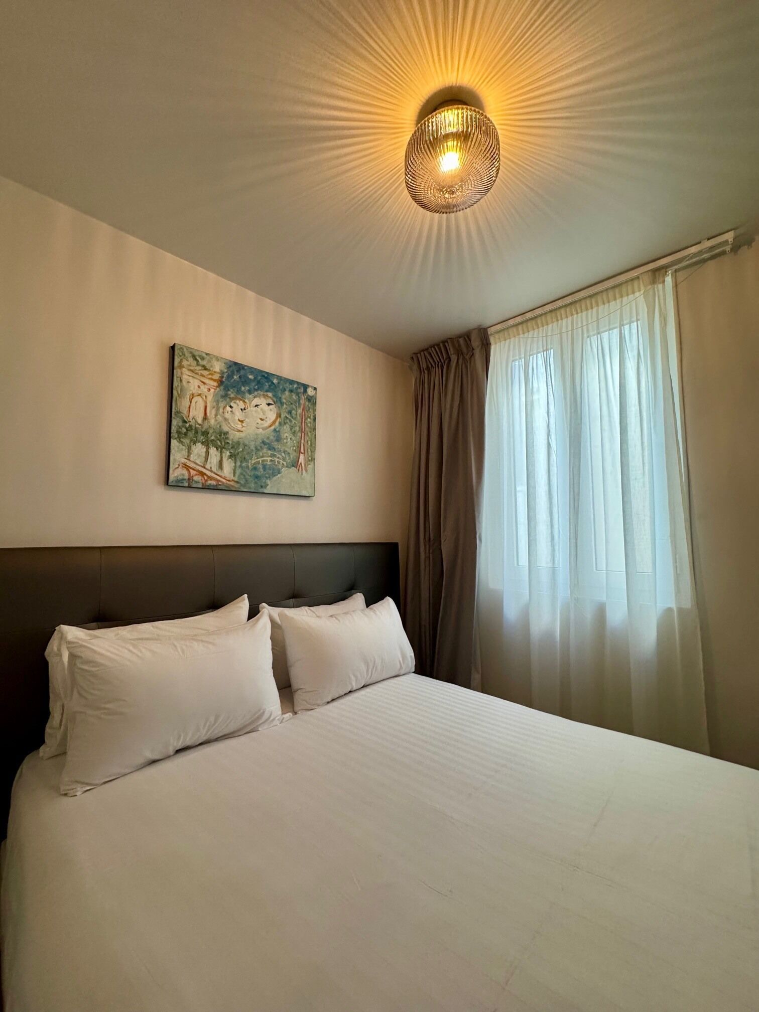 A hotel room with a bed, a painting on the wall, a ceiling light, and a window with curtains.