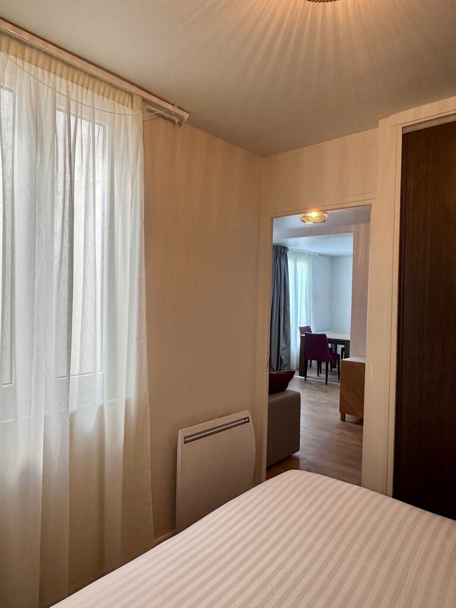 A hotel room with a bed, a radiator, a window with sheer curtains, and a view into a dining area with a table and chairs.