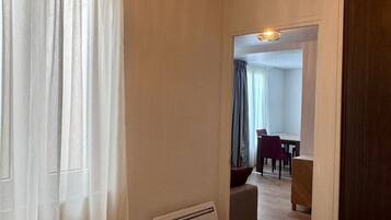 Een hotelkamer met een bed, een radiator, een raam met dunne gordijnen, en uitzicht op een eetgedeelte met een tafel en stoelen.