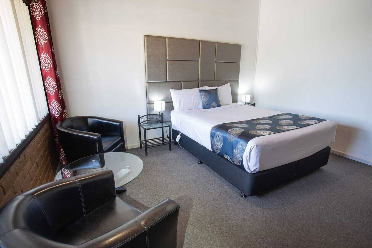 A hotel room with a bed, a chair, a small table, and a nightstand with a lamp.