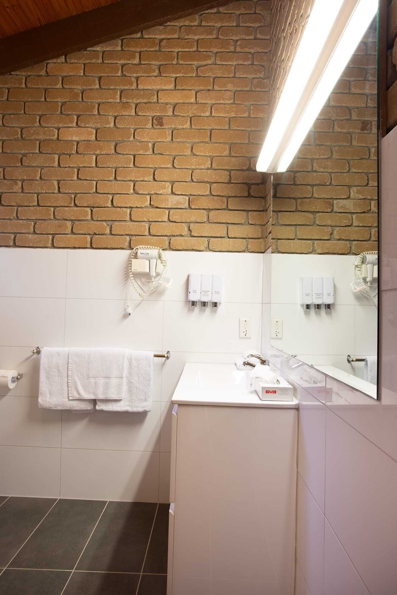 Casa de banho com parede de tijolos, azulejos brancos, espelho, lavatório e porta-toalhas.