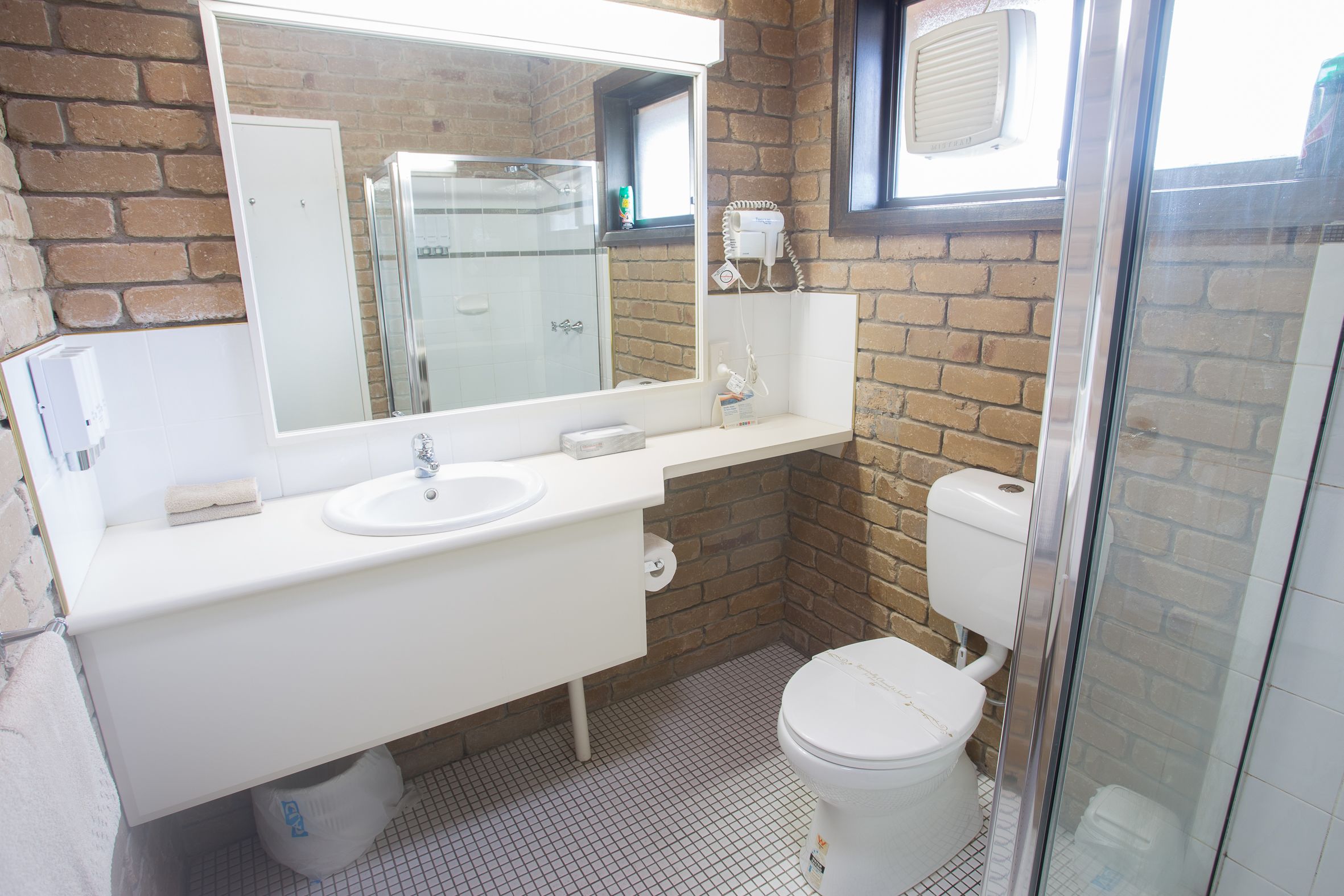 A bathroom with a white sink, mirror, and toilet, featuring a brick wall and a glass shower enclosure.