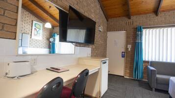 A compact room with a kitchenette, a flat-screen TV mounted on the wall, a small refrigerator, a desk with a chair, and a sofa.