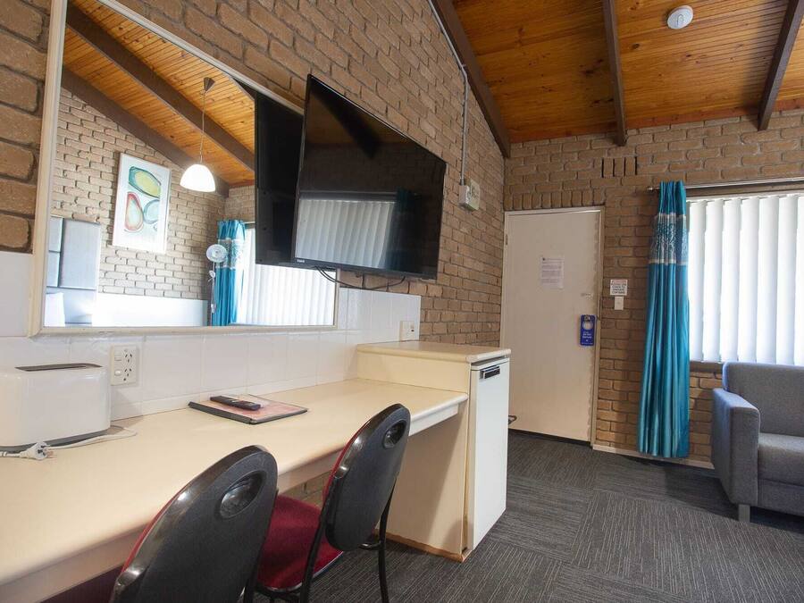 A compact room with a kitchenette, a flat-screen TV mounted on the wall, a small refrigerator, a desk with a chair, and a sofa.