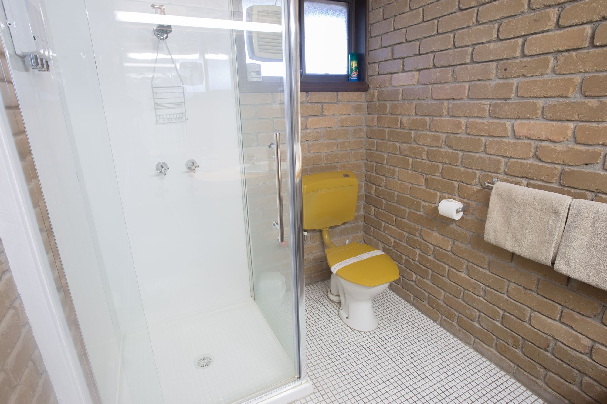 A bathroom with a glass shower enclosure, a yellow toilet, a brick wall, and a towel rack.