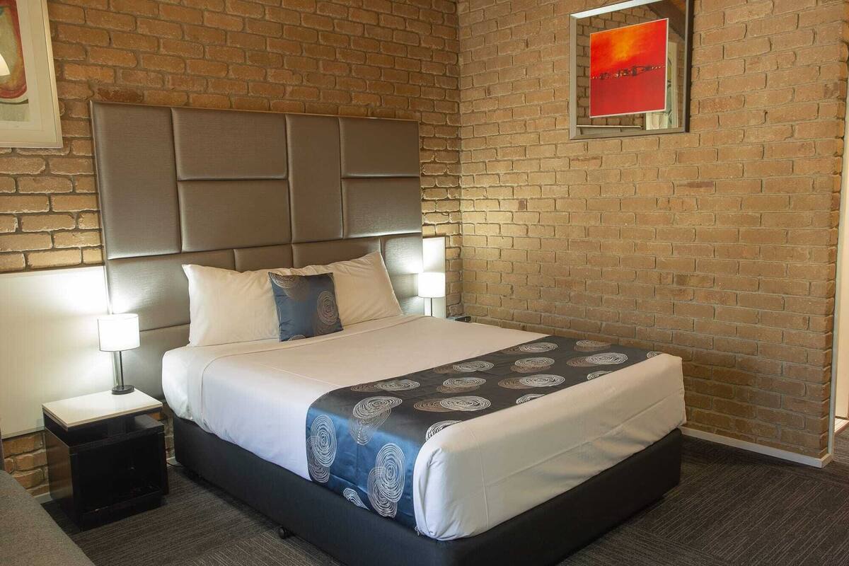 A hotel room with a brick wall, a large bed with a headboard, two bedside tables with lamps, and a framed picture on the wall.