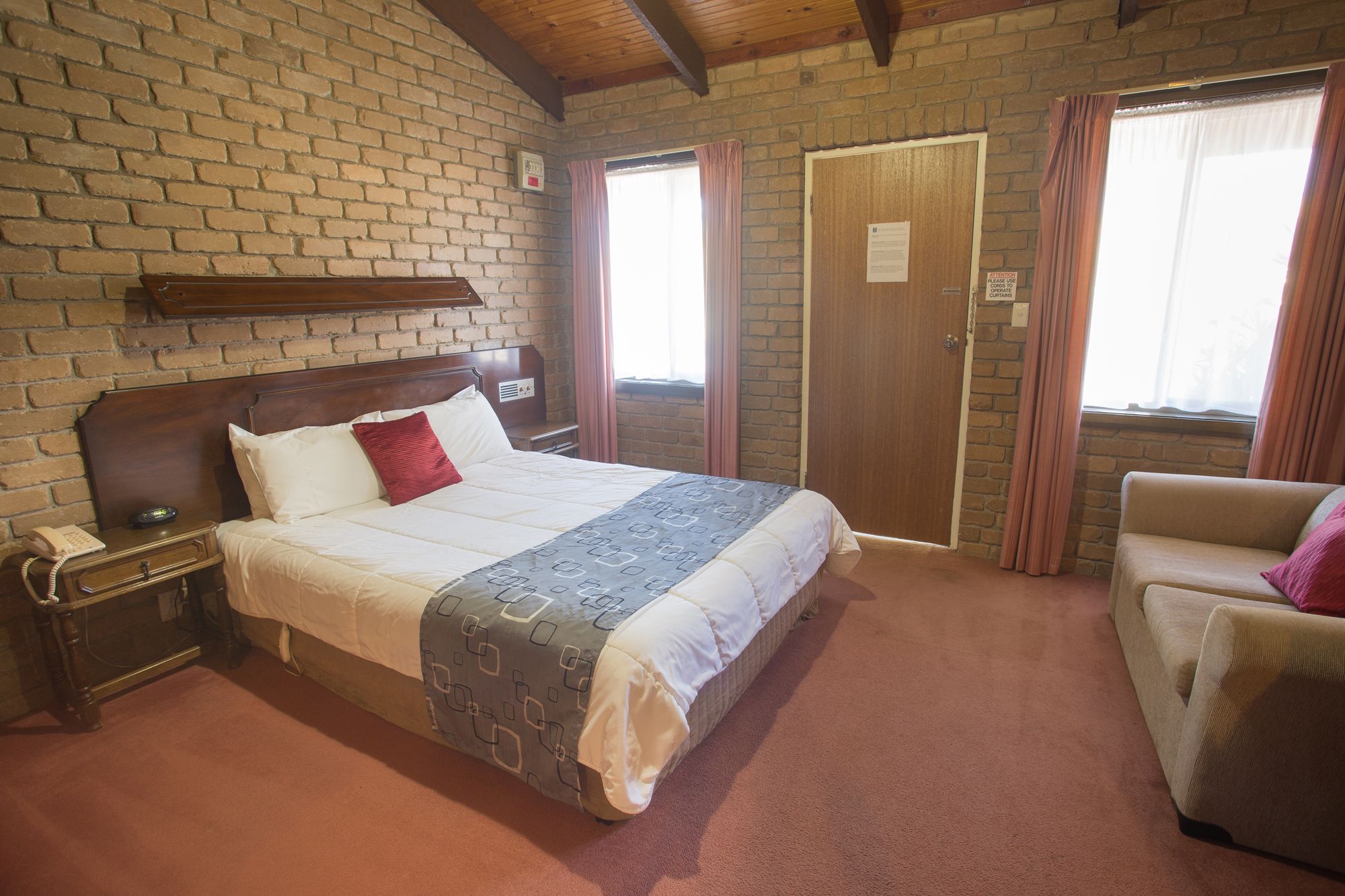 A hotel room with a brick wall, a wooden bed, a bedside table with a phone, a chair, and a door.