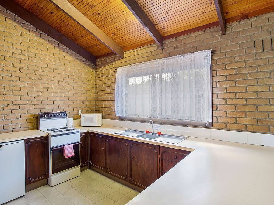 A kitchen with a brick wall, wooden ceiling beams, a white stove, a sink, and a window with a sheer curtain.