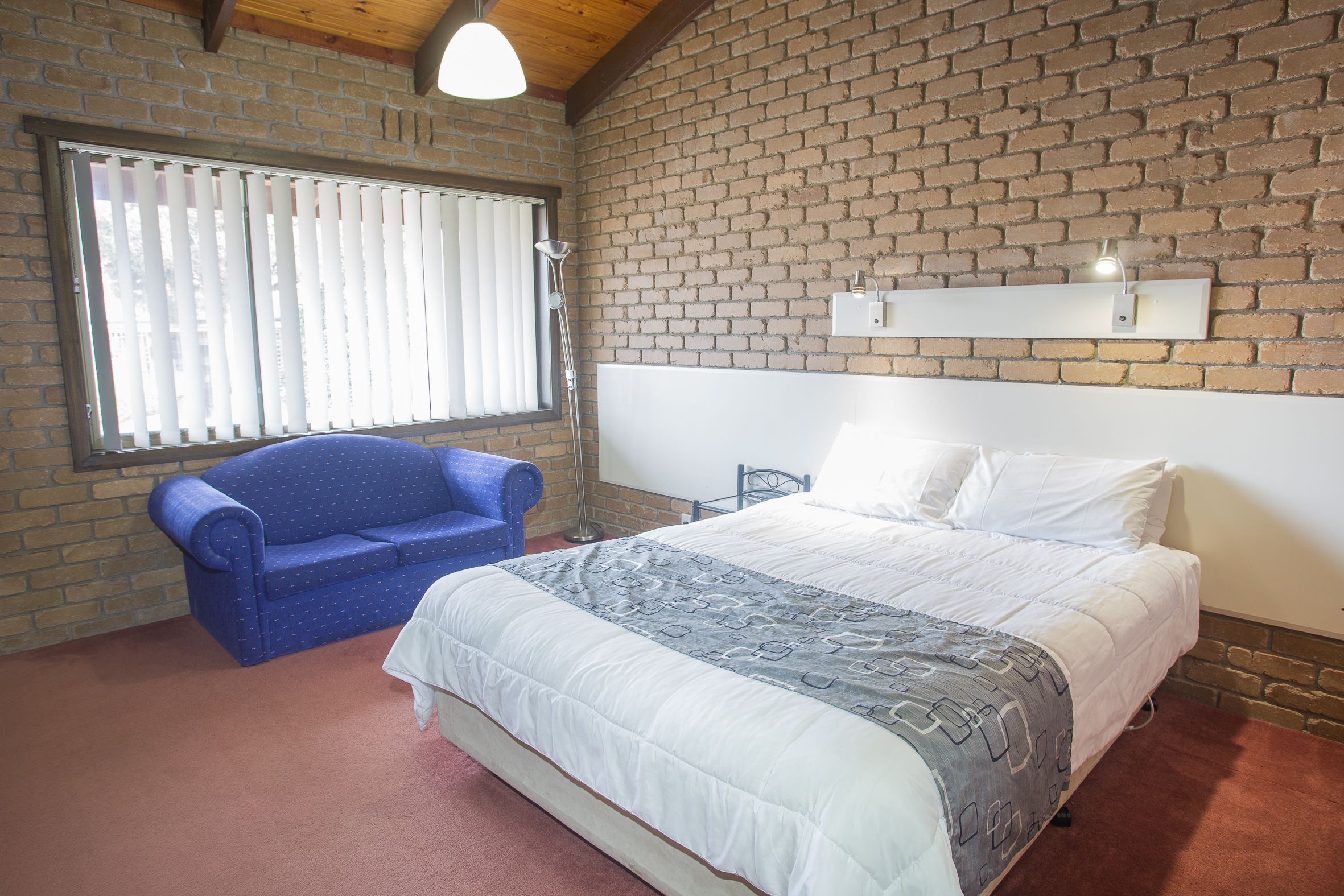 Una camera da letto con parete in mattoni, un letto con biancheria bianca, una sedia blu e una finestra con persiane.