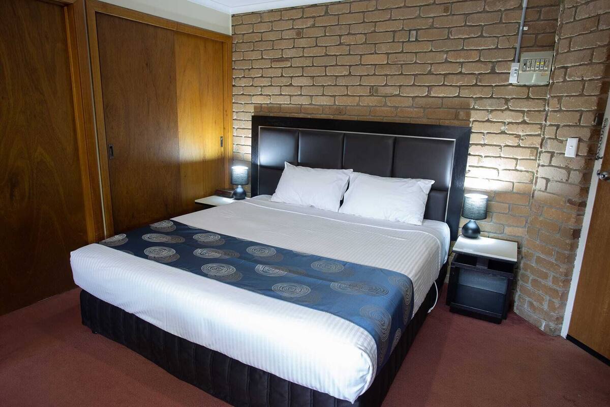 A hotel room with a bed, two bedside tables, a nightstand, a wooden wardrobe, and a brick wall.
