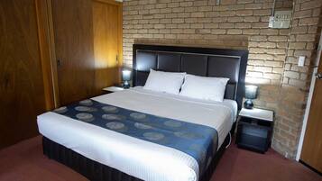 A hotel room with a bed, two bedside tables, a nightstand, a wooden wardrobe, and a brick wall.