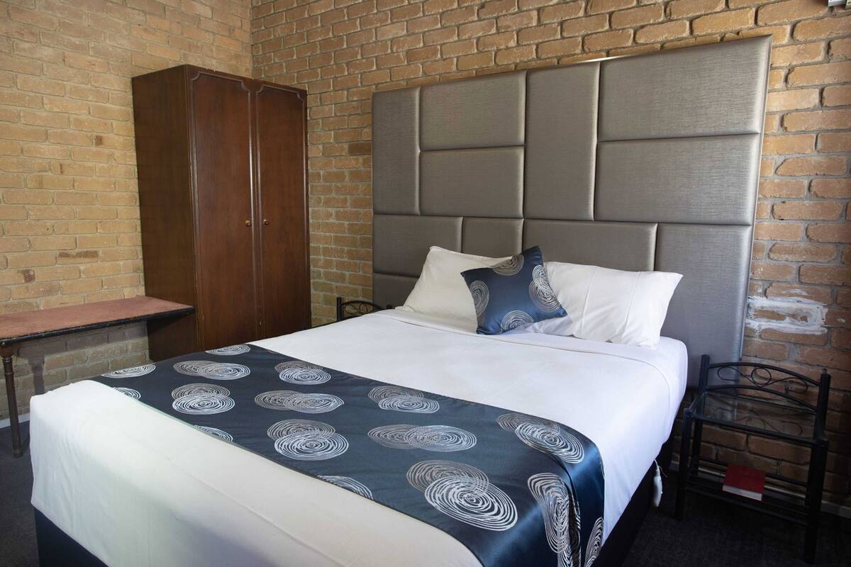 A hotel room with a brick wall, a wooden wardrobe, a bed with a grey headboard, and a bedside table.
