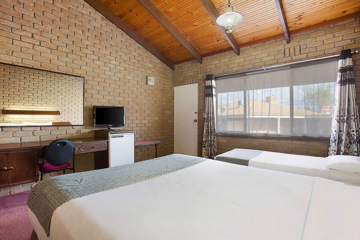 A hotel room with a brick wall, wooden ceiling beams, a desk with a television, two beds, and a window with curtains.