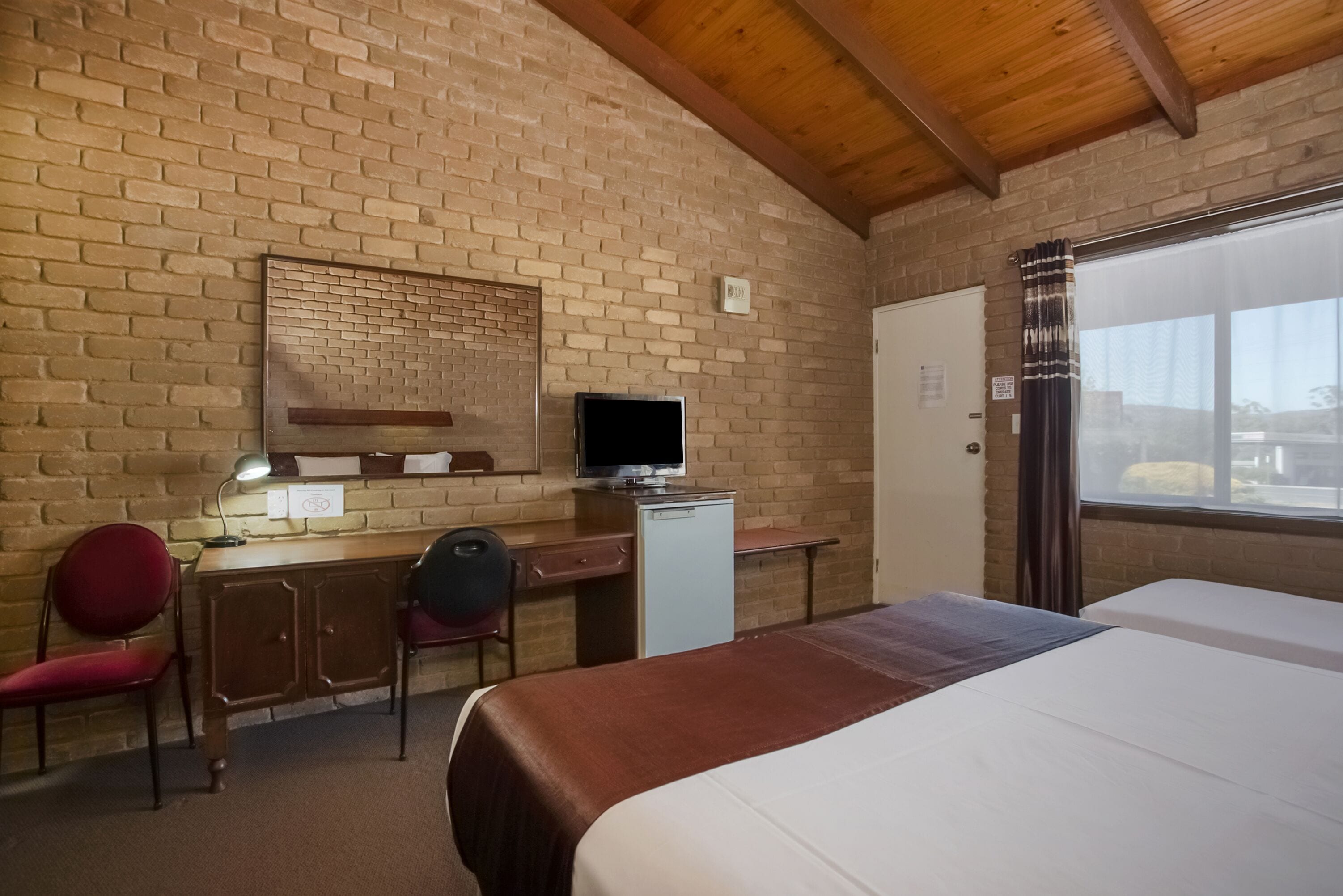 Quarto de hotel com parede de tijolos, teto de madeira, uma escrivaninha com computador, cama e janela com cortinas.