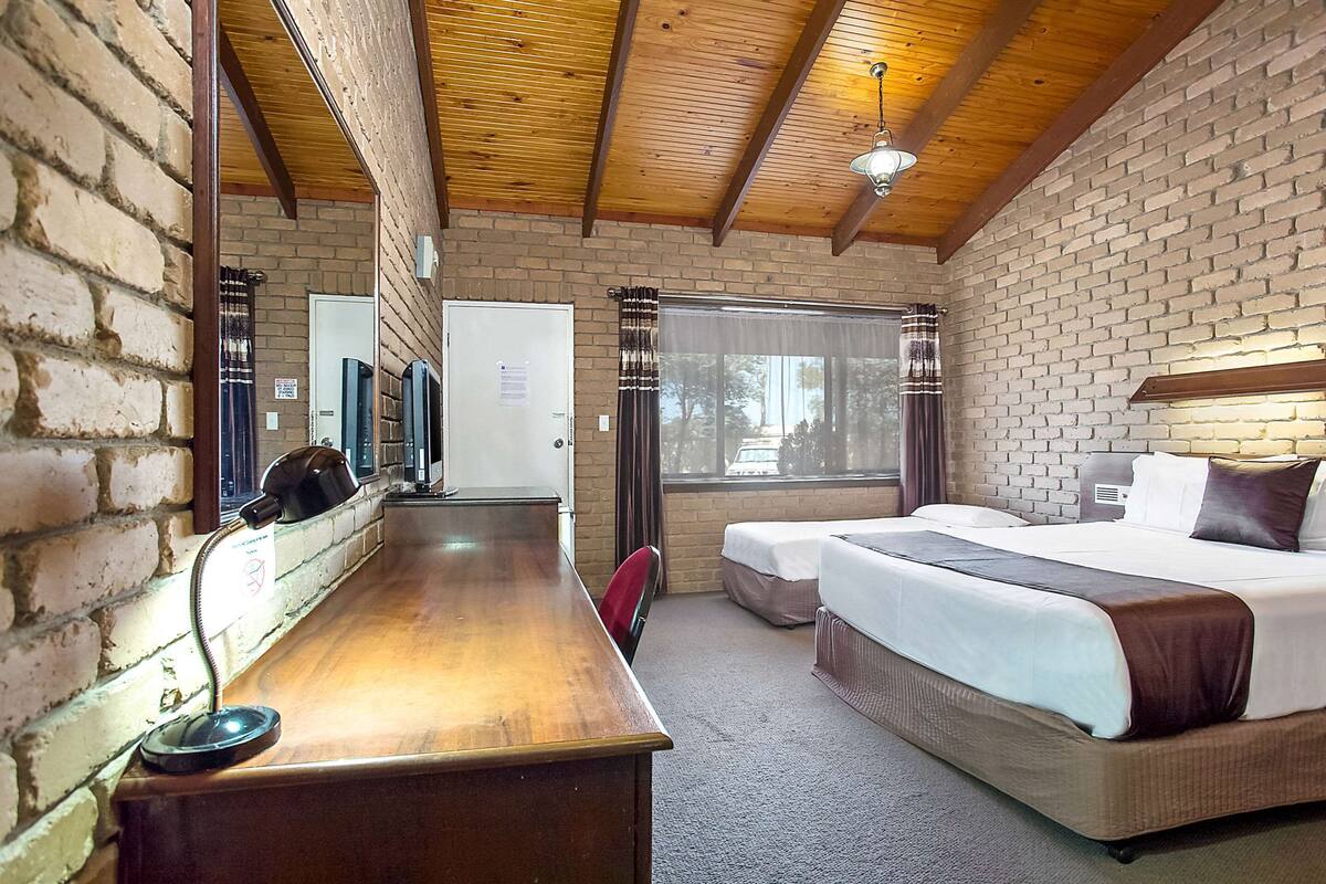 A hotel room with a wooden ceiling, brick walls, a desk with a telephone, a bed with a white and brown bedspread, and a window with curtains.