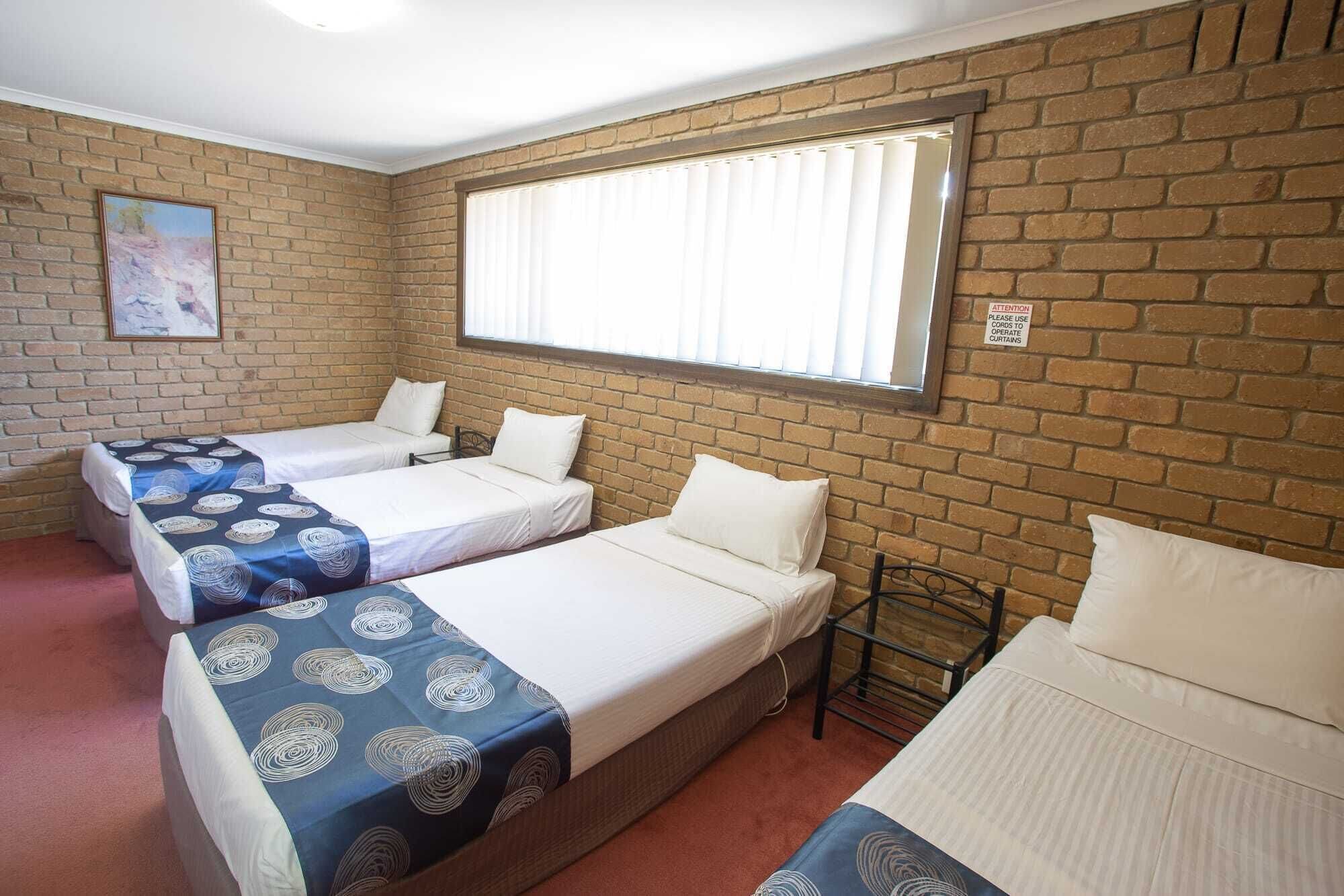 A room with three beds arranged in a row against a brick wall, a window with blinds, and a painting on the wall.